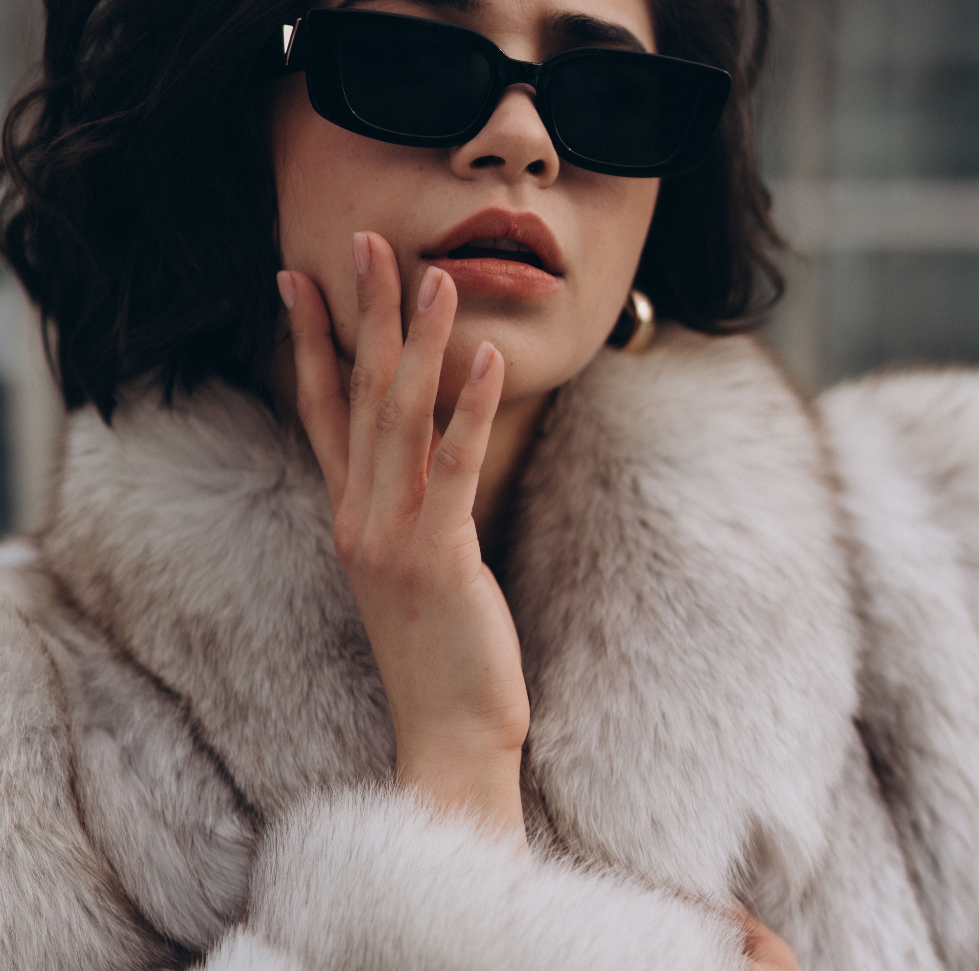 A woman wearing sunglasses and a fur coat touches her cheek