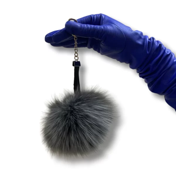 Blue leather glove wearing hand holds a grey fox pom pom keychain