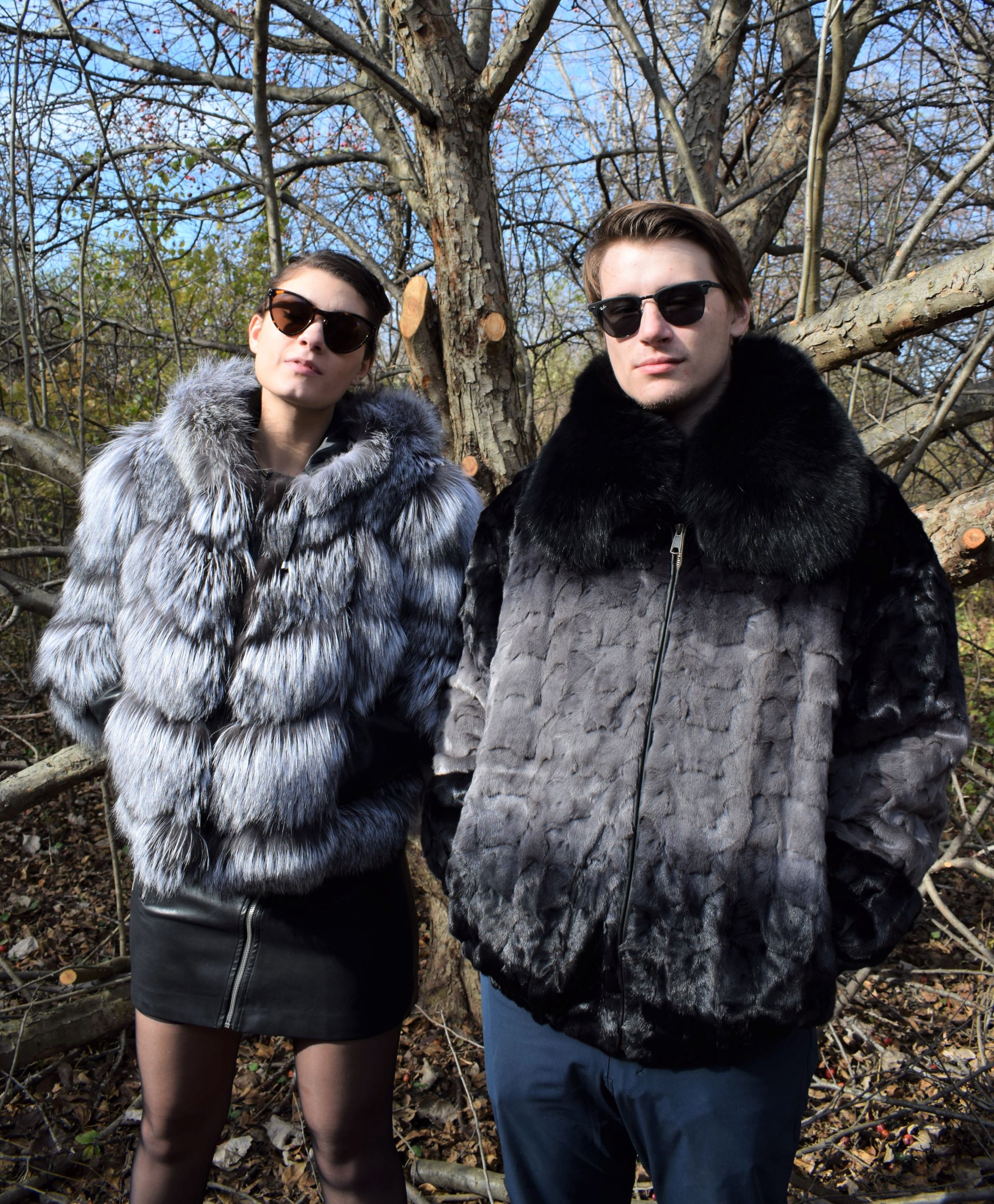 A male and female standing together in a wooded area while wearing black and grey fur jackets