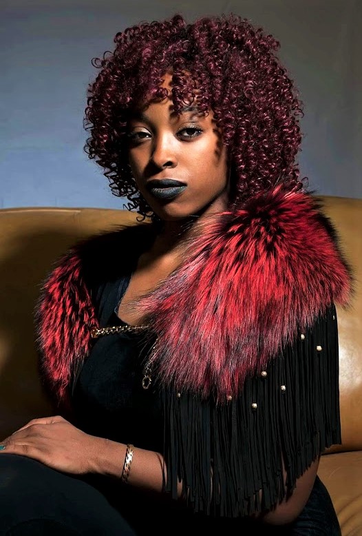 Woman sitting on a brown couch while wearing a black leather top with a red fur cowl